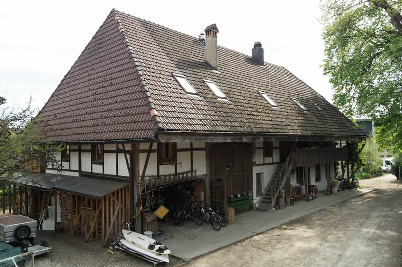 Two-story house with a steep, tiled roof and wooden beams, featuring a covered porch area with bicycles, a small boat, and a green bench.