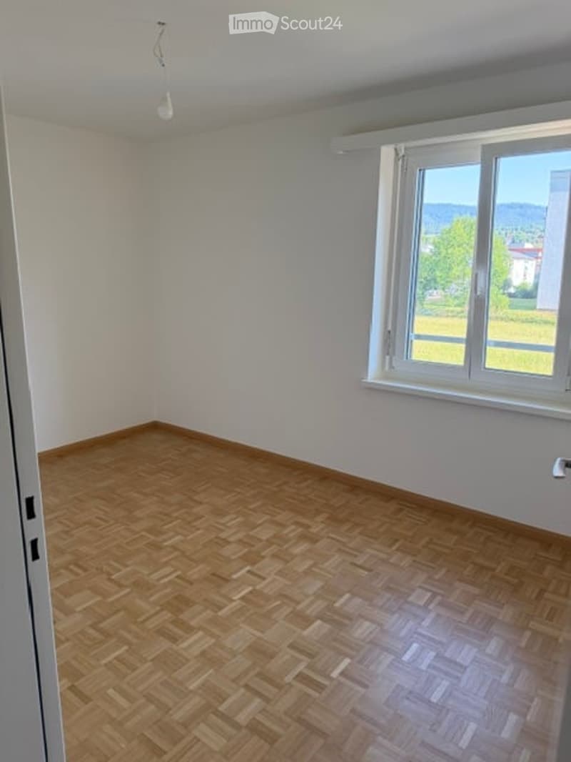 Empty room with wooden parquet flooring and white walls, featuring a large window with a view of greenery outside.