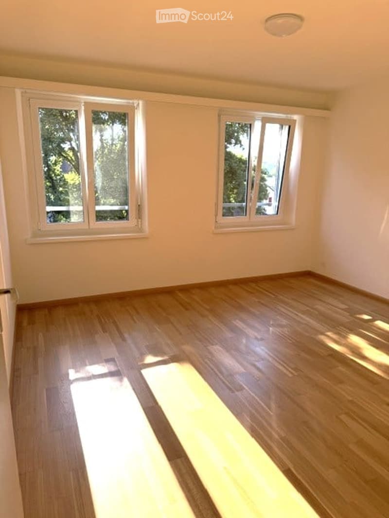 Empty room with wooden flooring and two windows, natural light coming through.
