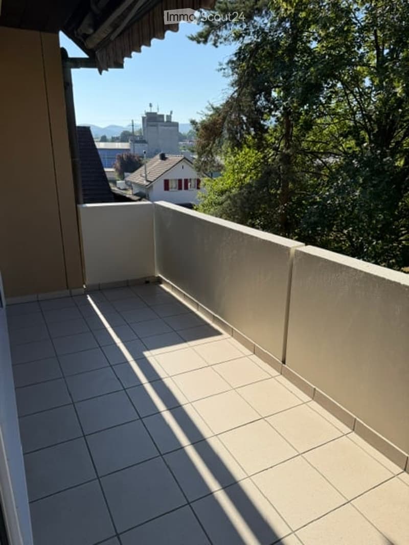 Balcony with tiled floor, white walls, and a view of trees and a house in the background.
