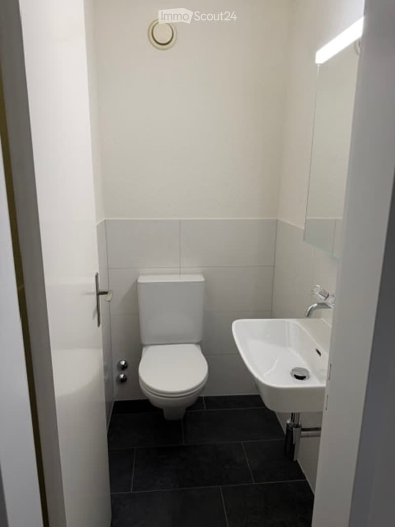 Modern, compact bathroom with white tiles, a toilet, a sink, and a mirror. The floor is dark grey tiles, and the walls are white.