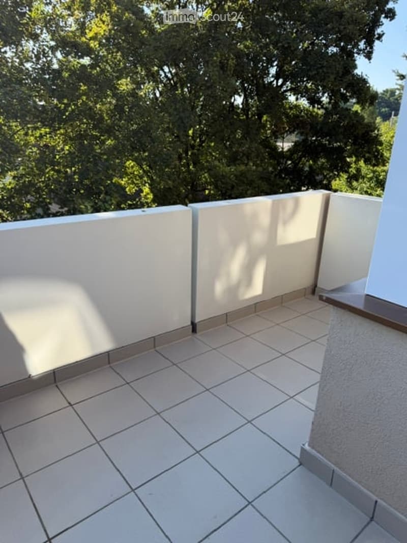 Balcony with white tiled floor and white walls, overlooking a lush green area with trees.