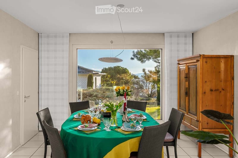 Dining room with a round table set for a meal, surrounded by wicker chairs, a large window with a view of the garden, and a wooden cabinet.