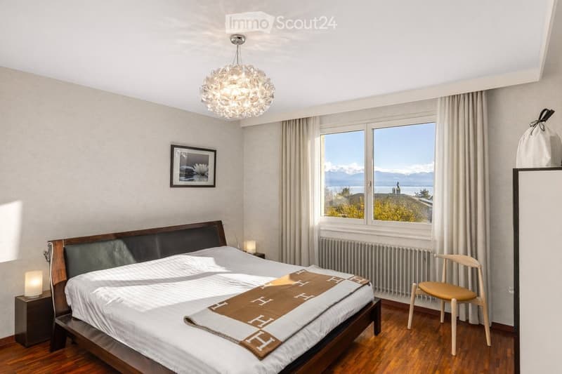 Modern bedroom with a large bed, wooden nightstand, chair, picture frame, and scenic view through large windows.