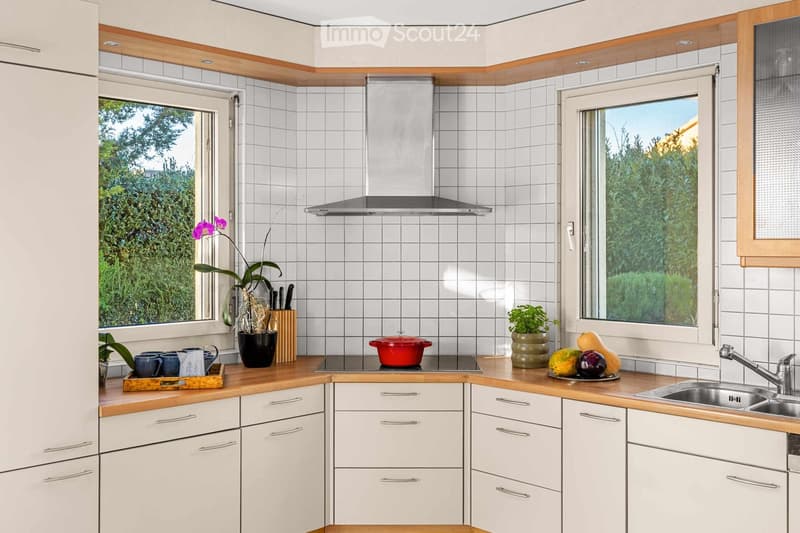 Modern kitchen with white tiled walls, wooden countertops, and white cabinets. Features a stainless steel sink, stove, and various kitchen items including cups, a pot, and a fruit bowl.