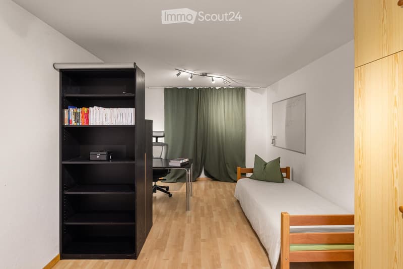 A small, modern bedroom with a single bed, a wooden desk, a bookshelf, and a curtained window. The room features light wooden flooring and white walls.