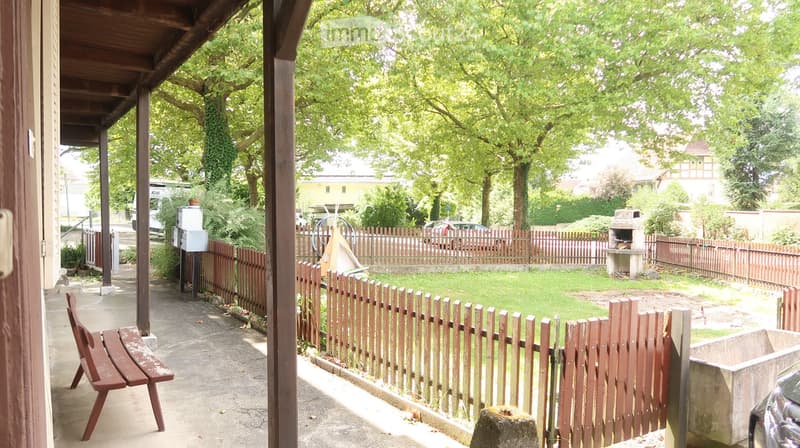 covered porch, wooden bench, wooden fence, backyard with grass and small brick barbecue, trees, and garden shed, car parked nearby