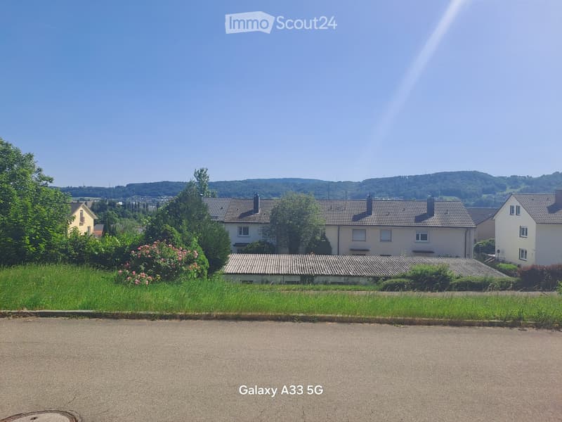 Residential houses with tiled roofs, surrounded by greenery and trees, with a clear blue sky and distant hills in the background.