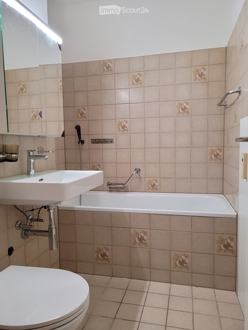 Bathroom with beige tiled walls and floor, featuring a white sink, bathtub, toilet, and a mirror above the sink. A handrail is attached to the wall next to the bathtub.