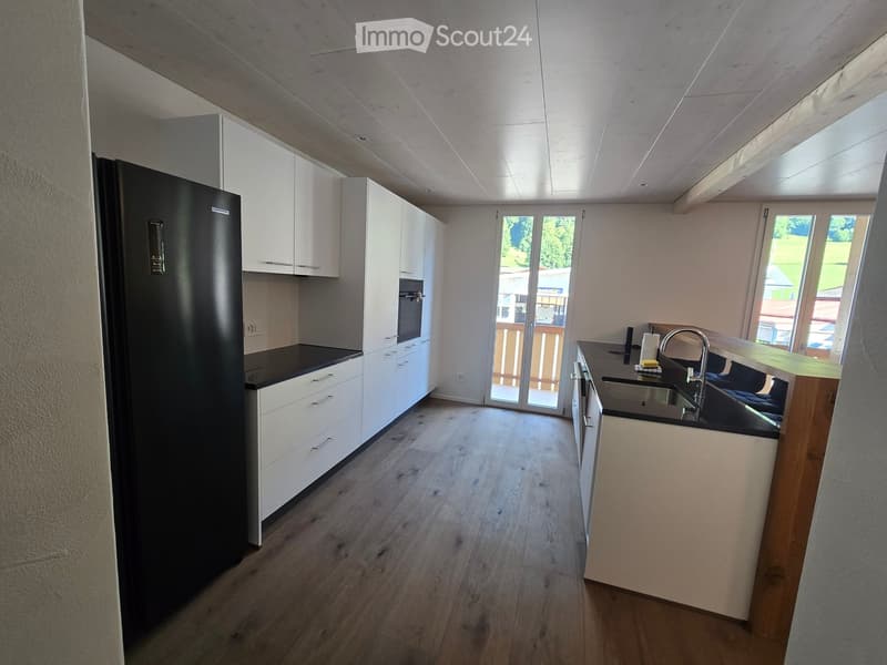 Modern kitchen with white cabinets, black countertops, a stainless steel sink, and a refrigerator. The space features wooden flooring and large windows with a view of the outdoors.