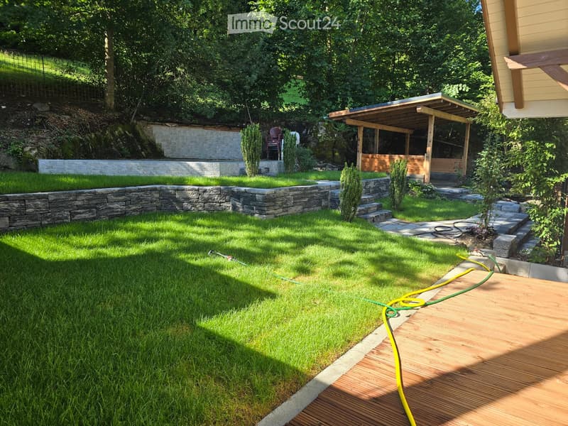 Well-maintained garden with a wooden deck, green lawn, stone walls, and a covered outdoor seating area.