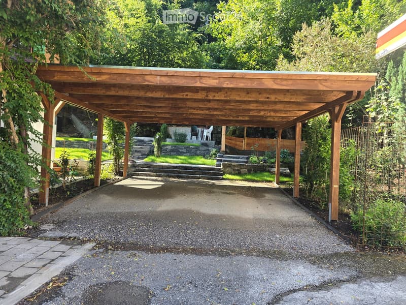 Wooden carport with a gravel driveway, surrounded by greenery and a stone staircase leading to a garden area.