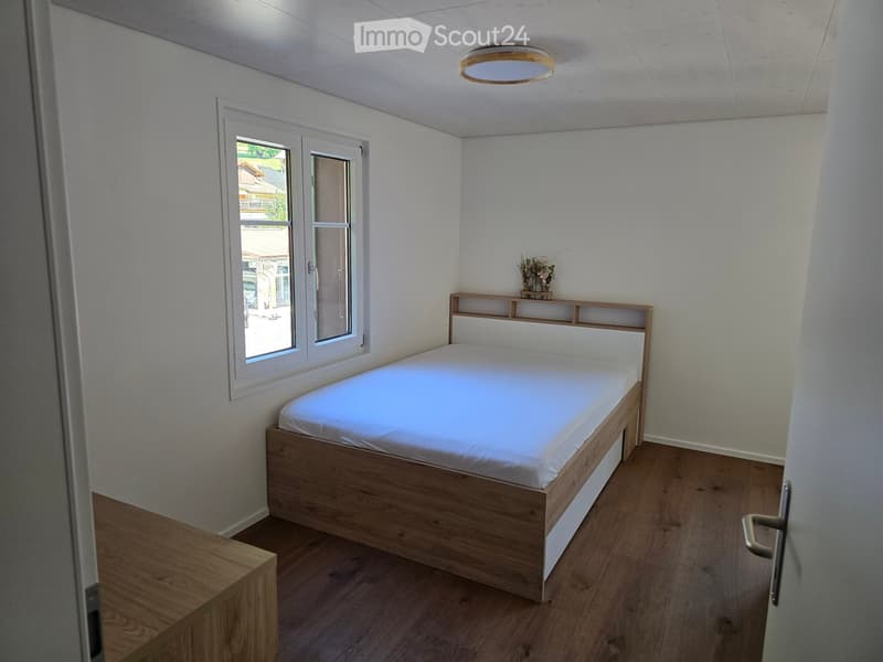 A small, modern bedroom with a wooden bed, white bedding, a headboard with built-in shelves, and a vase on the nightstand. The room features light-colored walls, wooden flooring, and a window with white frames.