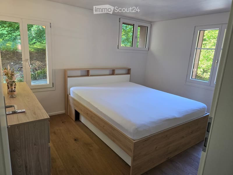 A small, well-lit bedroom with a wooden bed frame, white mattress, nightstand, and vase on the nightstand. The room has light-colored walls and wooden flooring.