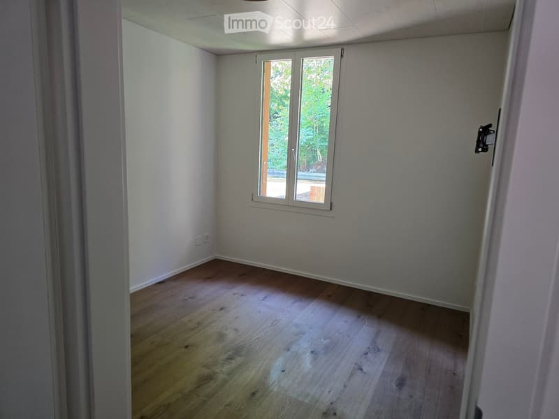 Empty room with light wooden flooring, white walls, and a large window with white frames, allowing natural light to enter.