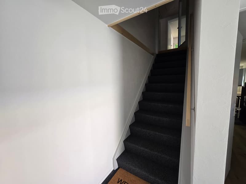 A modern staircase with dark carpeted steps and white walls, featuring a wooden handrail and a partially open door leading to another room.