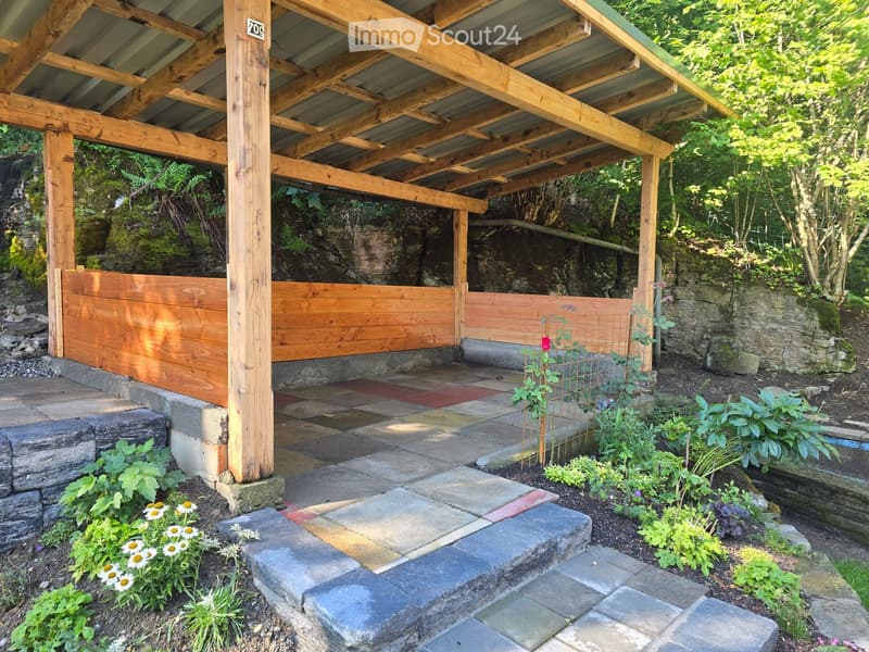 A wooden pergola with a stone patio, surrounded by lush greenery and flowers, including daisies and climbing plants, with a stone wall and steps leading up.