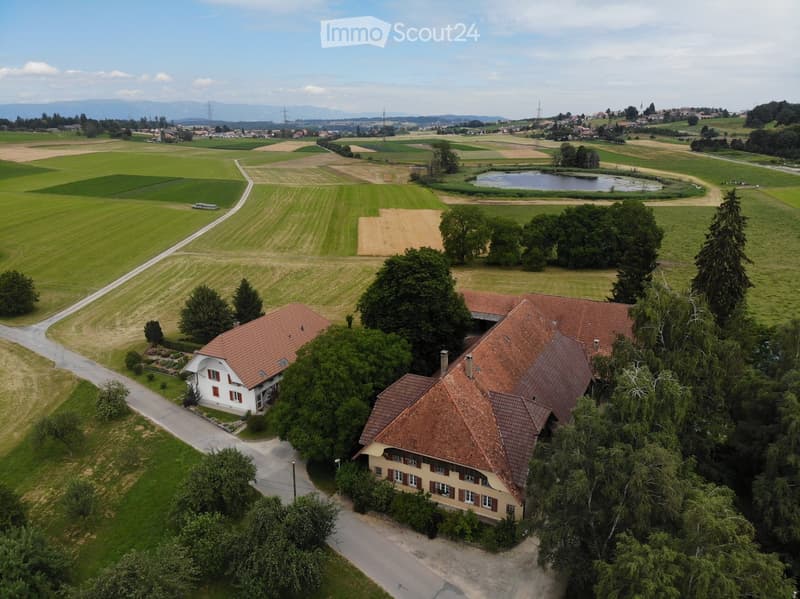 Aerial view of a two-story house with red-tiled roofs, surrounded by trees and fields. The house has multiple windows and is situated near a driveway. The surrounding area includes agricultural fields, a small pond, and distant mountains.