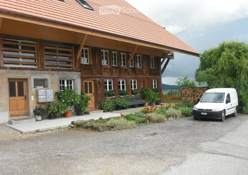 Two-story house with wooden facade and stone base, featuring multiple windows, a balcony, and a well-maintained garden with potted plants and a bench.