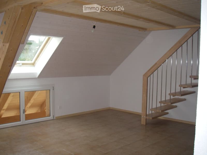 Empty attic space with wooden ceiling beams, skylight window, tiled floor, and a wooden staircase leading upwards.