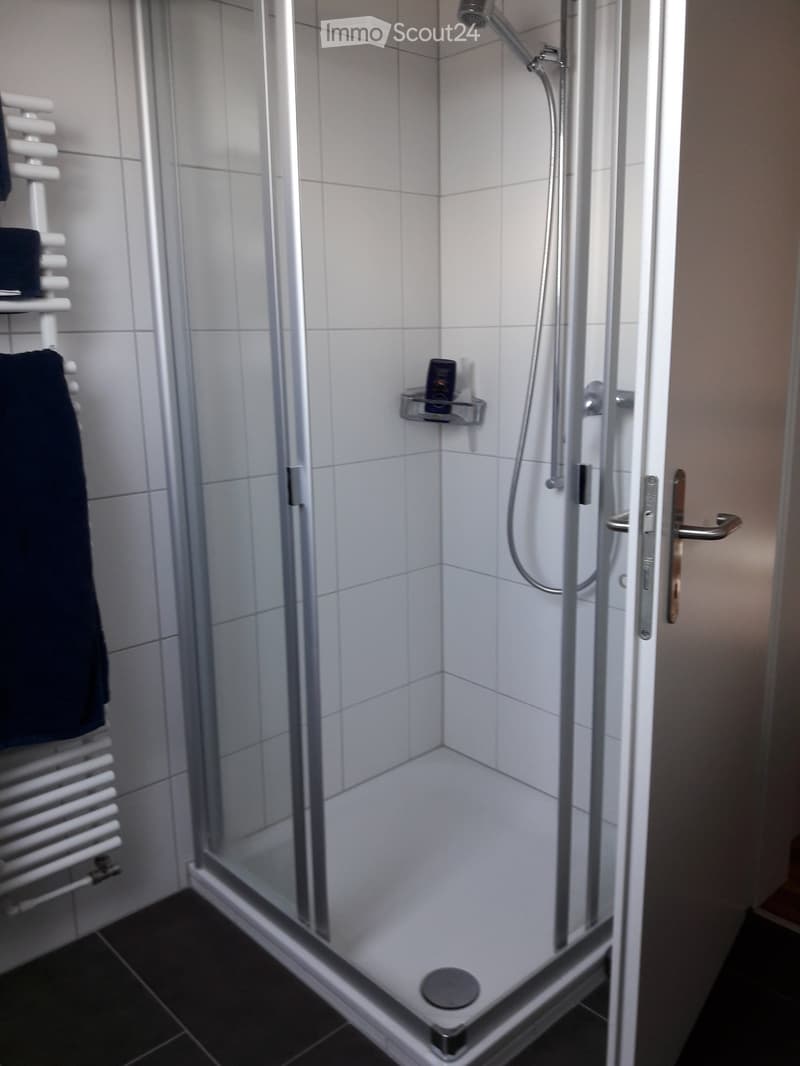 Modern bathroom with a glass-enclosed shower, white tiled walls, a towel rack with a dark towel, and a small shelf inside the shower holding toiletries.