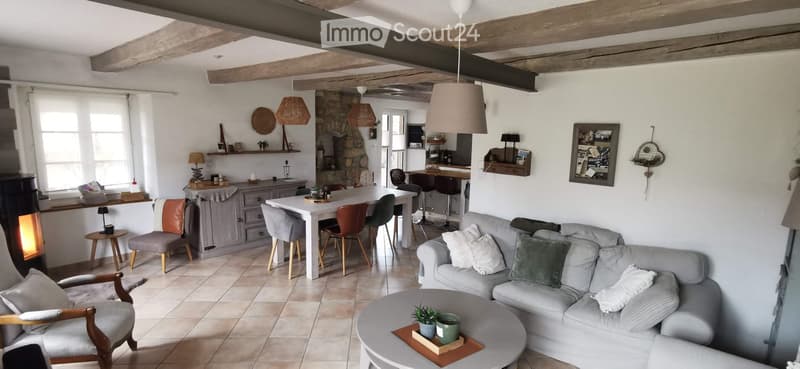 Open-plan living and dining area with exposed wooden beams, a fireplace, a grey sofa, a coffee table, and a dining table with chairs.