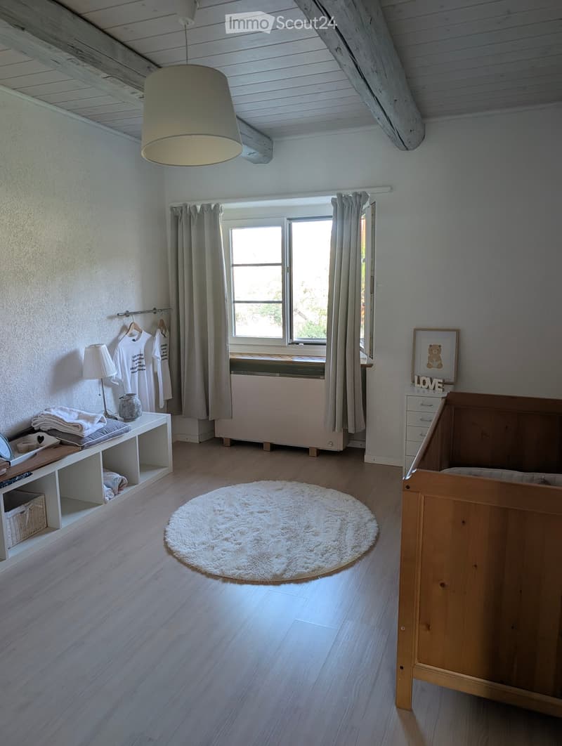 Bright bedroom with wooden flooring, a round white rug, a wooden bed, a small table with a lamp, and a window with curtains.
