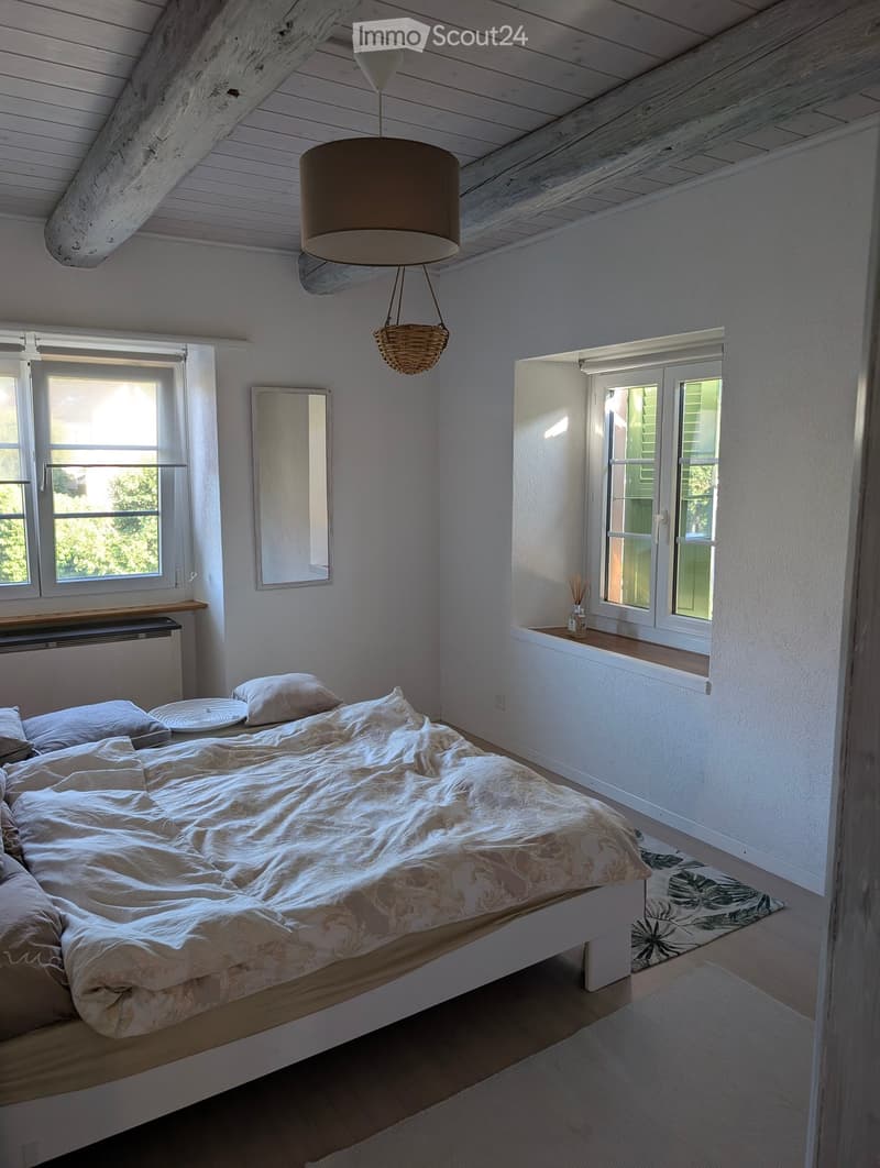 Cozy attic bedroom with wooden slanted ceilings, white walls, large windows, a bed with white bedding, a mirror, and a pendant light.