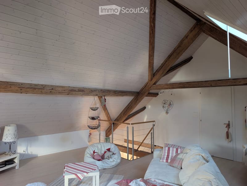 Bright living room with exposed wooden beams, white walls, and a staircase leading to an upper level. Features a white sofa with striped pillows, a bean bag chair, a small coffee table, and a floor lamp.
