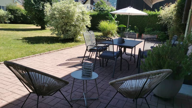 A well-maintained garden patio with a brick floor, featuring a dining table with chairs, a small round table with a stool, and an umbrella providing shade. The area is surrounded by greenery and a grassy lawn.