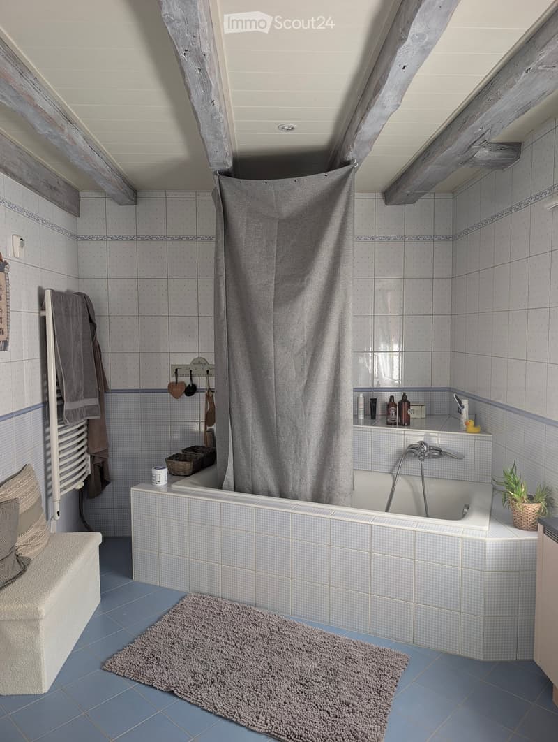 Cozy bathroom with blue tiles, a bathtub, a shower curtain, a towel rack with towels, a small plant, and various toiletries on the edge of the tub.