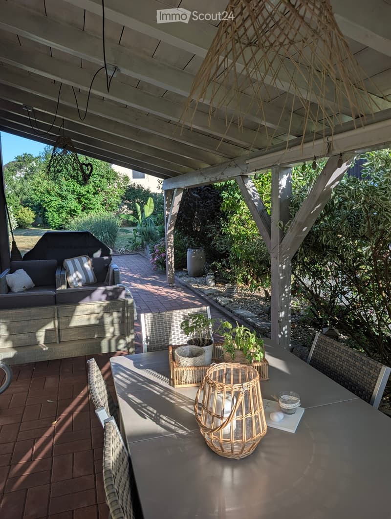 A covered outdoor patio with a wooden ceiling, a sofa with cushions, a dining table with chairs, and decorative items including a wicker basket and candle holder. The area is surrounded by greenery and has a brick pathway leading to a garden.