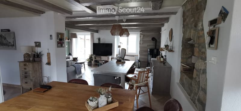 Bright living and dining area with exposed wooden beams, a sofa, coffee table, dining table with chairs, television, and decorative items.