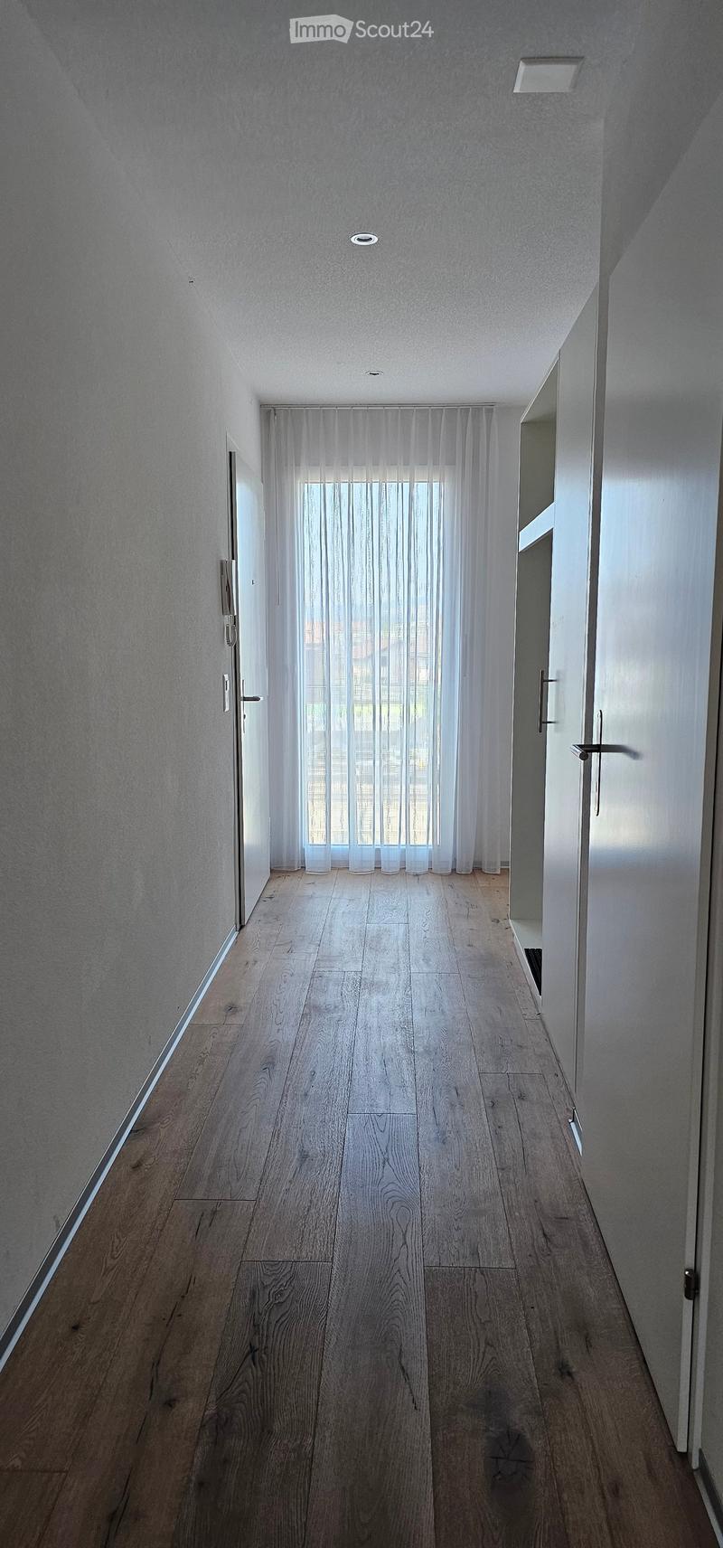 A modern, minimalist hallway with light wooden flooring, white walls, and recessed ceiling lighting. Features a large window with sheer white curtains and several doors along the sides.