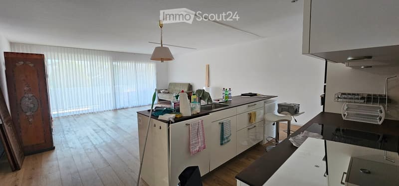 A modern kitchen with white cabinets, a central island, and a sink. There is a sofa in the background, a curtain covering a large window, and various kitchen items on the counter.