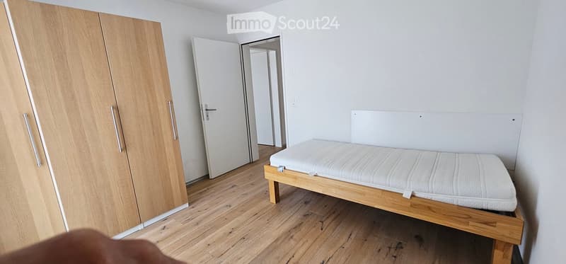 Minimalist bedroom with a wooden bed frame and mattress, a large wooden wardrobe, and white walls.