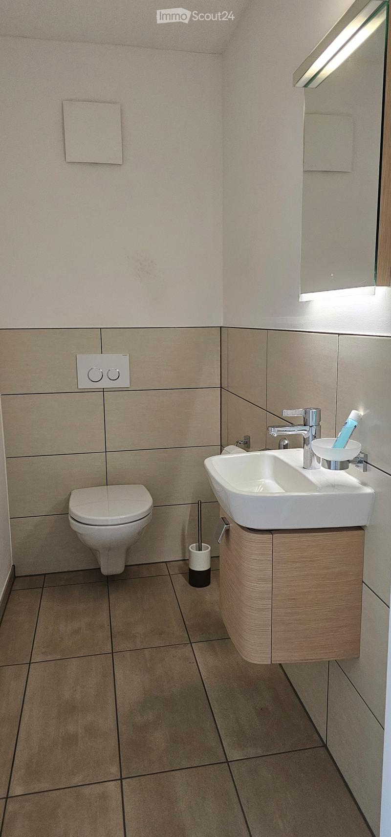 Modern bathroom with a white toilet, a sink with a modern faucet, a toothbrush holder, and a mirror with built-in lighting. The walls are tiled in a light beige color, and the floor is tiled in a darker beige.