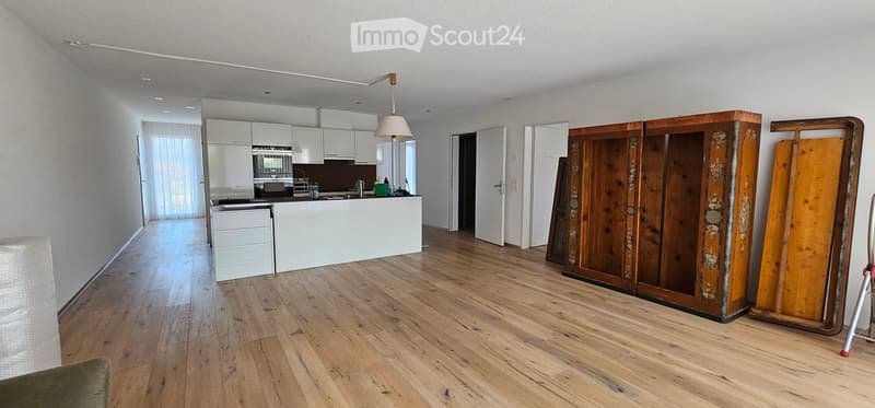 Open-plan living and kitchen area with wooden flooring, white kitchen cabinets, a central island, and a hanging lamp. A large wooden wardrobe is positioned against the wall.