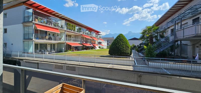 Modern multi-story residential building with balconies, red awnings, and a scenic mountain view in the background.