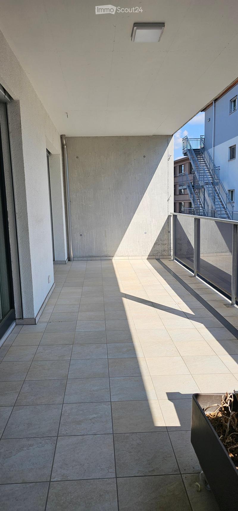 Modern balcony with tiled floor, concrete walls, and metal railings. Sunlight casts shadows on the floor, and a small planter is visible on the right side.