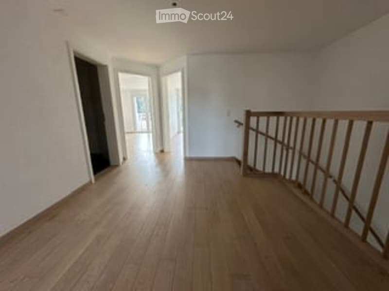 Empty room with wooden flooring and white walls, featuring a wooden staircase railing and open doorway leading to another room.