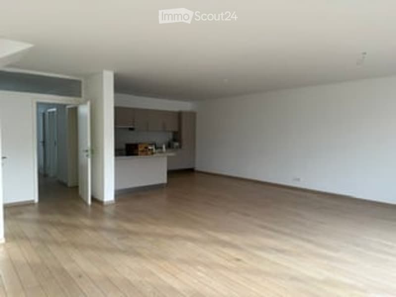 Empty room with wooden flooring, white walls, and a small kitchen area visible in the background.