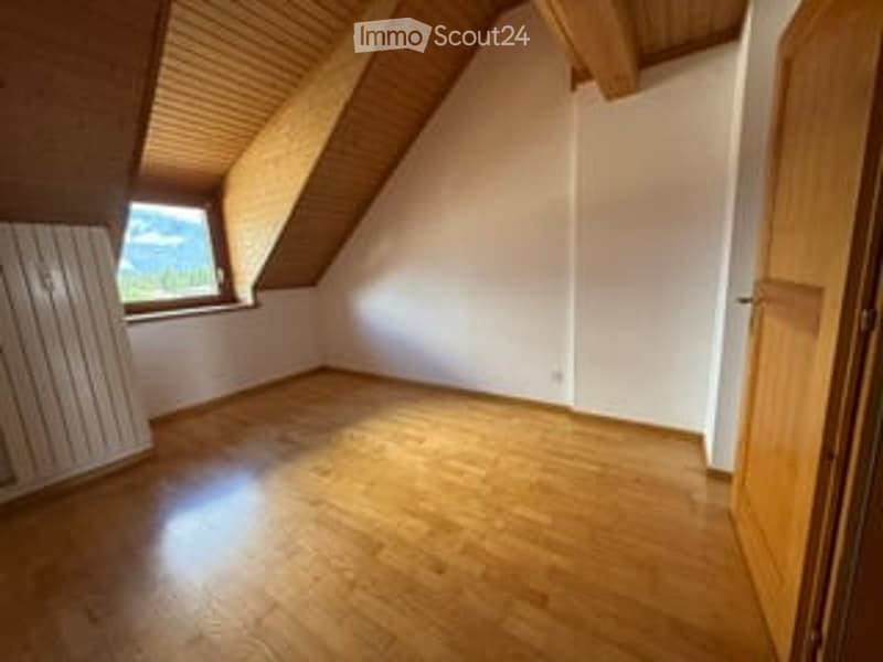 Empty attic room with wooden floor, white walls, exposed wooden beams, and a single window with a view of the landscape.