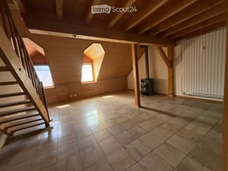 Empty attic space with wooden beams, tiled floor, and exposed brick walls. Features a wooden staircase leading up, a small window, and a radiator on the wall.