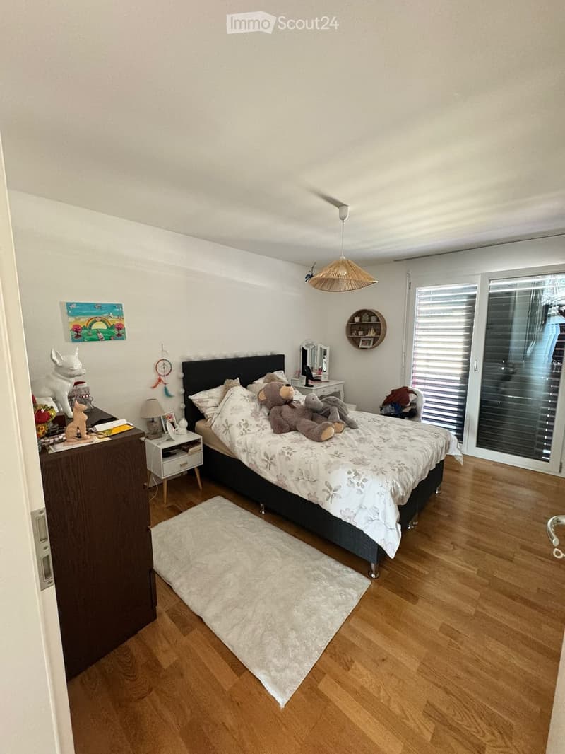 Cozy bedroom with wooden flooring, a large bed with a floral duvet, a nightstand with a lamp, a mirror, and decorative items including a stuffed animal and artwork on the walls.