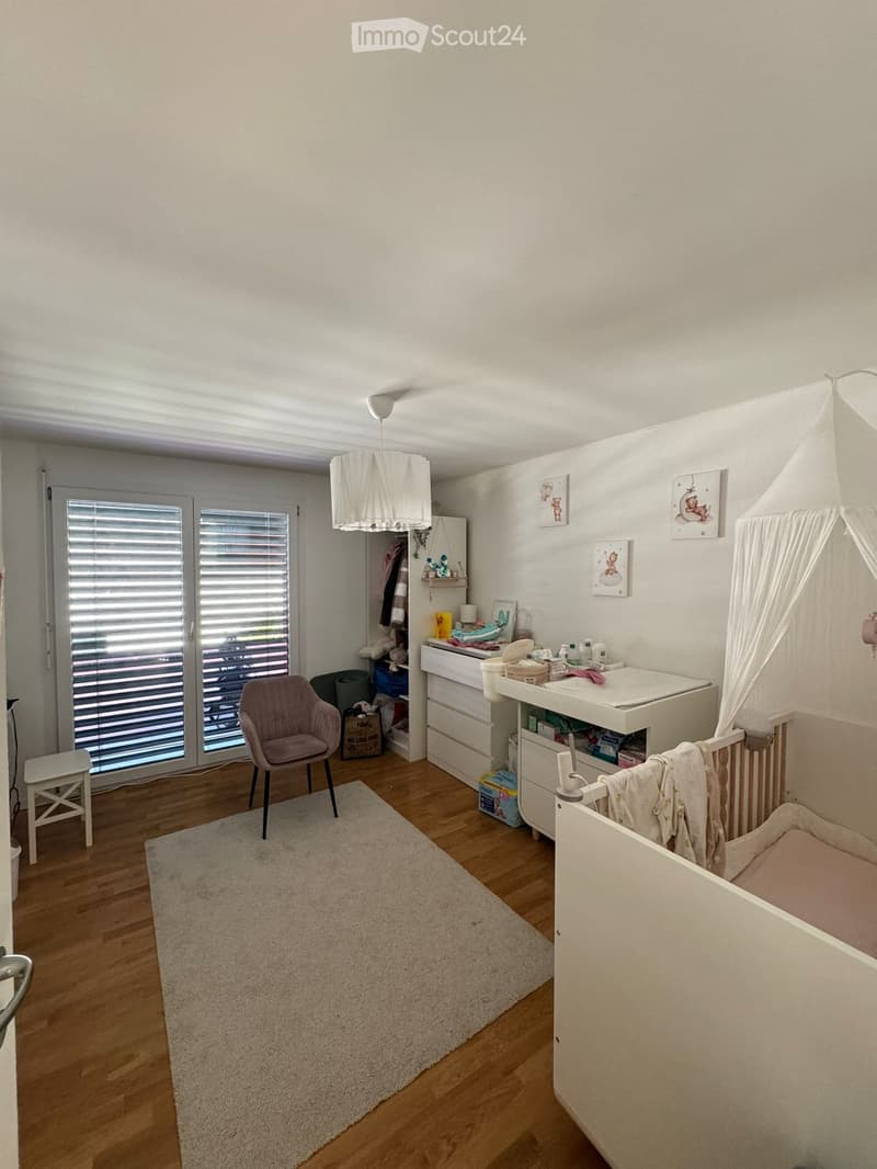 Brightly lit nursery with wooden flooring, a baby crib, changing table, and a cozy chair near the window.
