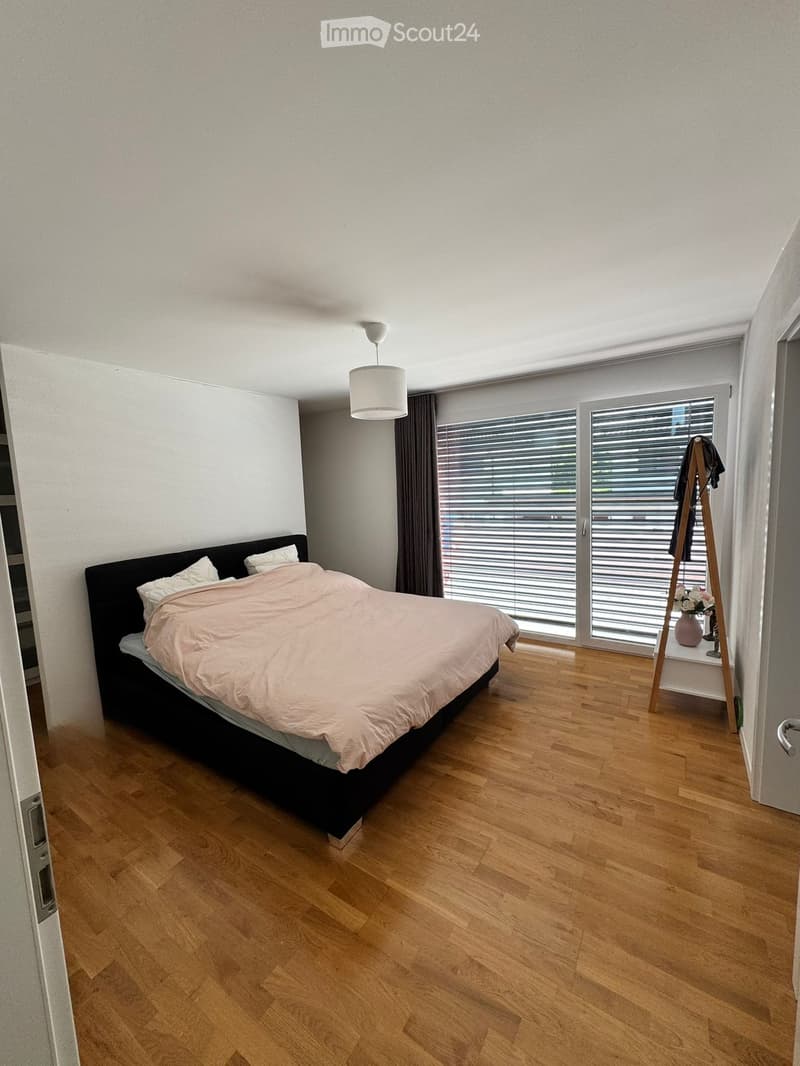 Modern bedroom with wooden flooring, large window with blinds, bed with beige bedding, pendant light, and coat rack with a vase on the shelf.