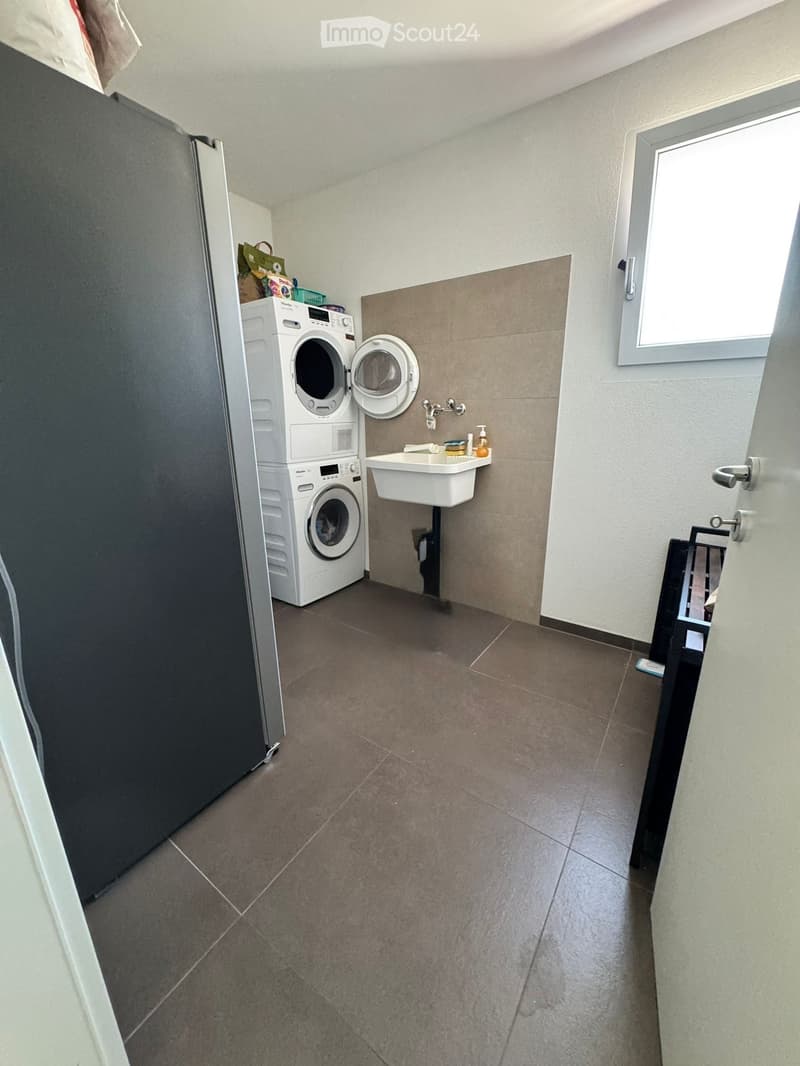 A small bathroom with a washing machine, a sink, and a window. The floor is tiled, and the walls are painted white with a beige accent wall.
