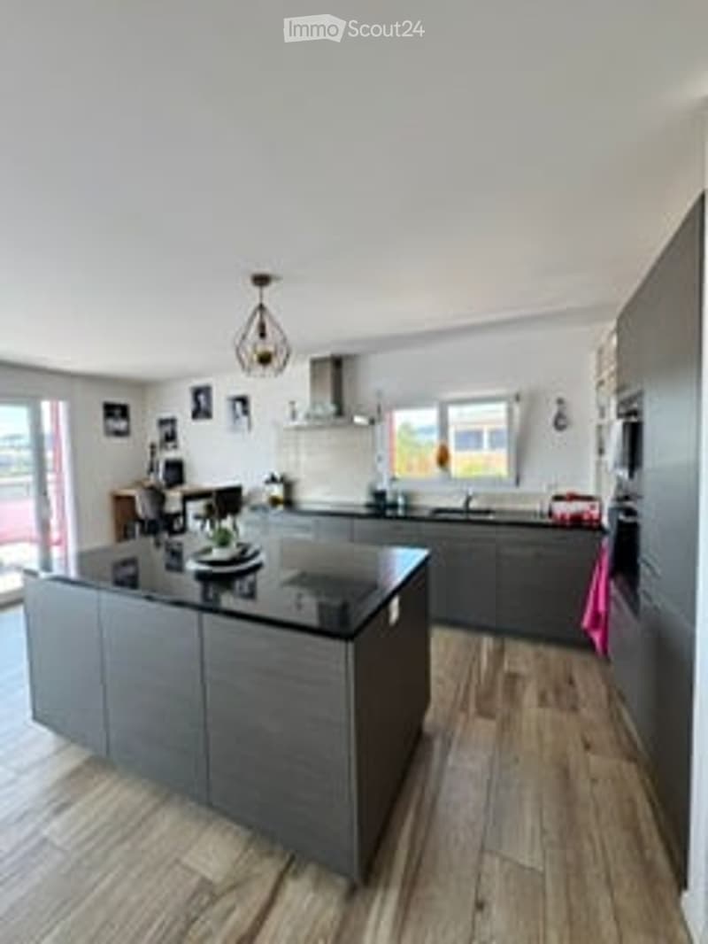 Modern kitchen with grey cabinets, a central island, and wooden flooring. Features a sink, faucet, and a hanging light fixture.