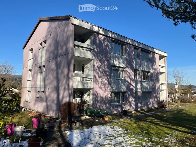 Modern two-story building with balconies, surrounded by a small garden area with potted plants and outdoor furniture.